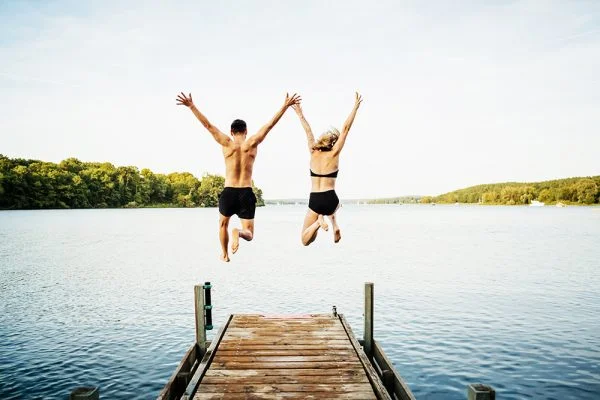 two happy-looking people in swimsuits jumping off dock into lake
